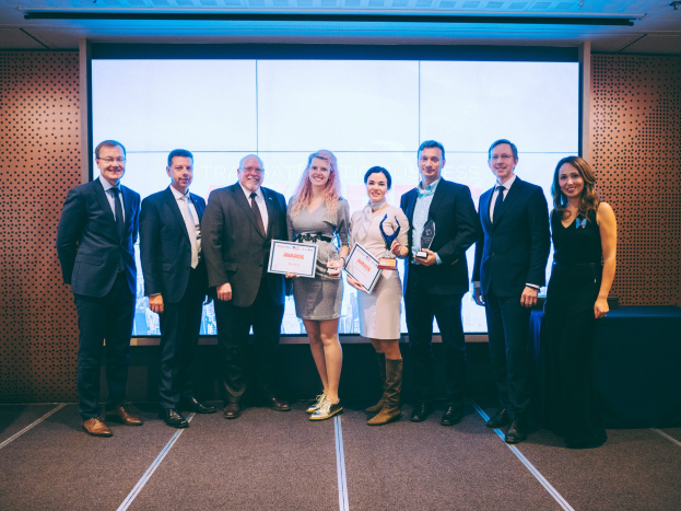 Gruppe von lächelnden Menschen mit Urkunden, die vor einem Bildschirm mit einem Tisch und einem Gegenstand im Hintergrund stehen, Gewinner der Internationalen Business Awards 2018.
