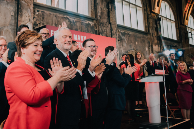 Eine Gruppe von Menschen, die vor einem Publikum klatschen, mit einem Podium, einem Mikrofon und einer Texttafel rechts und Stühlen, einer Wand, Fenstern und Lichtern im Hintergrund.