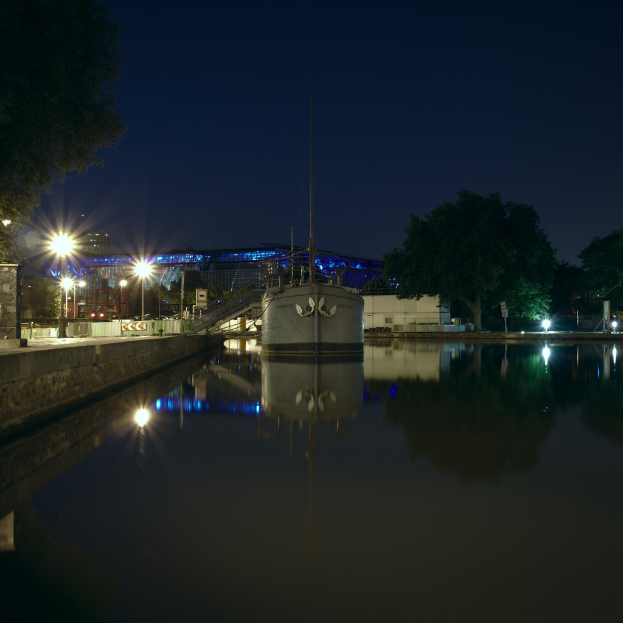 Mehrere Gebäude, ein Tank und Spiegelungen im Wasser mit Treppen, Straßenlaternen, Bäumen und dem Himmel im Hintergrund.