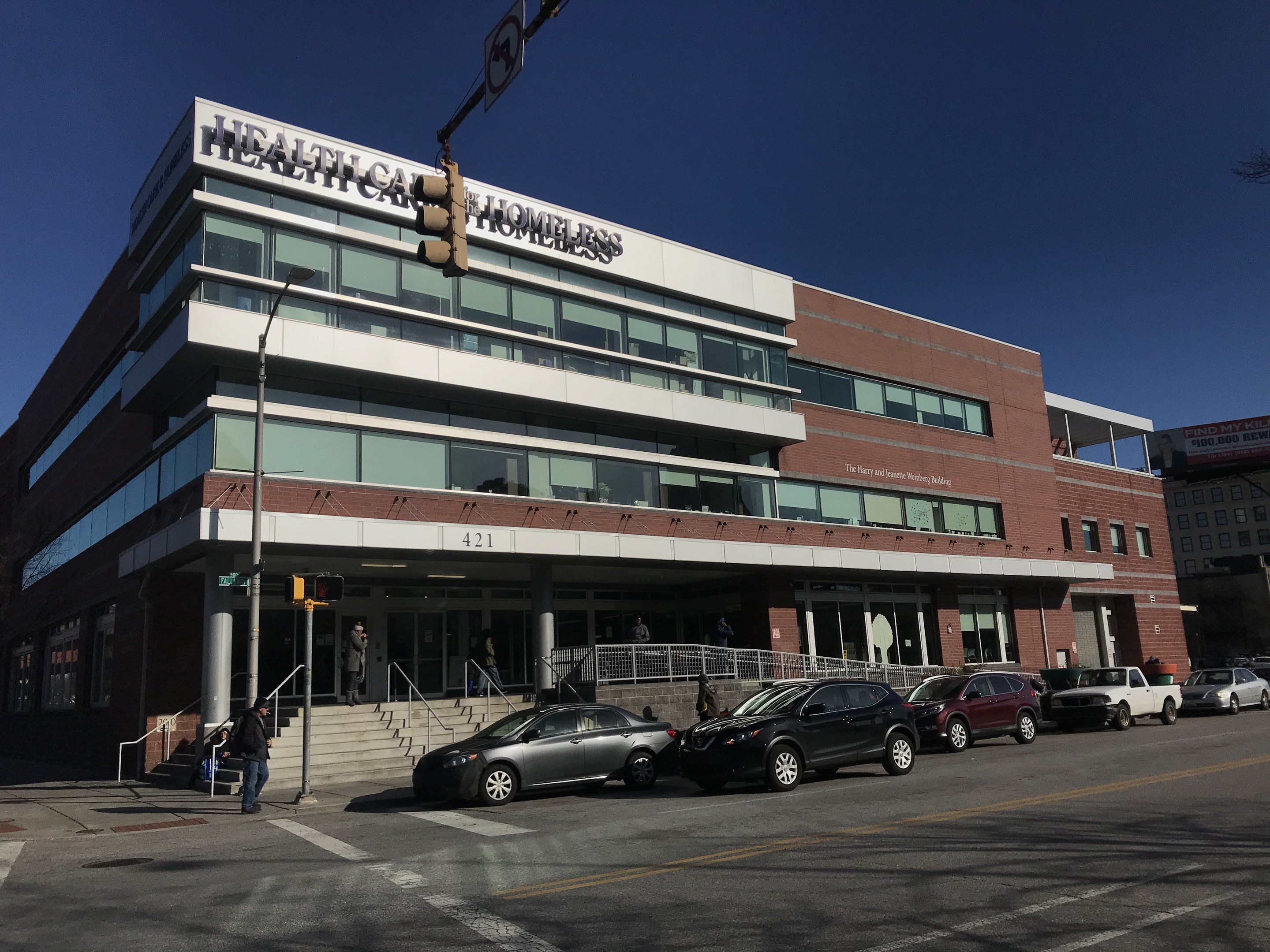 Großes Gebäude mit der Aufschrift 'New York City Health Care Homeless Center' mit parkenden Autos davor, umgeben von Straßeninfrastruktur, Fußgängern auf dem Gehweg und Bäumen unter einem klaren blauen Himmel.