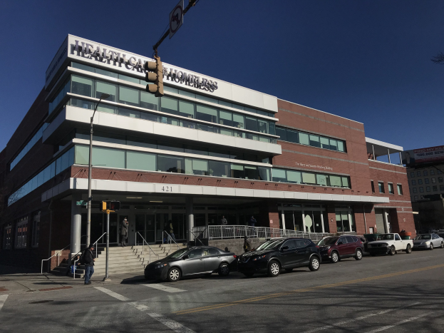 Großes Gebäude mit der Aufschrift 'New York City Health Care Homeless Center' mit parkenden Autos davor, umgeben von Straßeninfrastruktur, Fußgängern auf dem Gehweg und Bäumen unter einem klaren blauen Himmel.