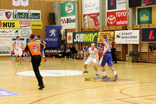 Ein Basketballfeld mit Spielern im Vordergrund und einer hölzernen Wand im Hintergrund, die mit Plakaten und Bannern geschmückt ist.
