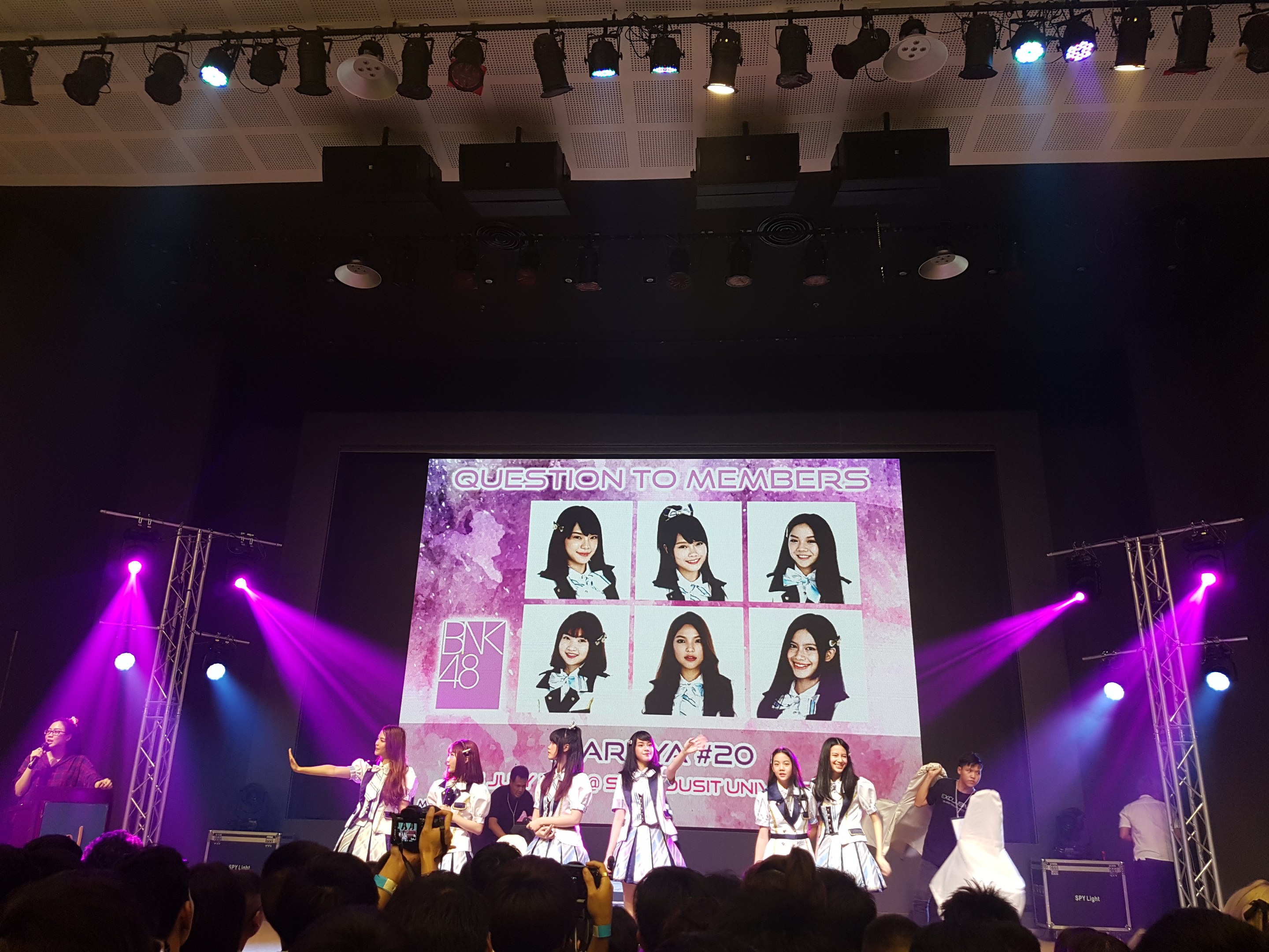 Eine Gruppe von Menschen performs auf einer hell erleuchteten Bühne mit Trägern, während eine Menge darunter die Veranstaltung aufzeichnet; ein großer Bildschirm im Hintergrund zeigt Bilder der K-Pop-Gruppe BK48.