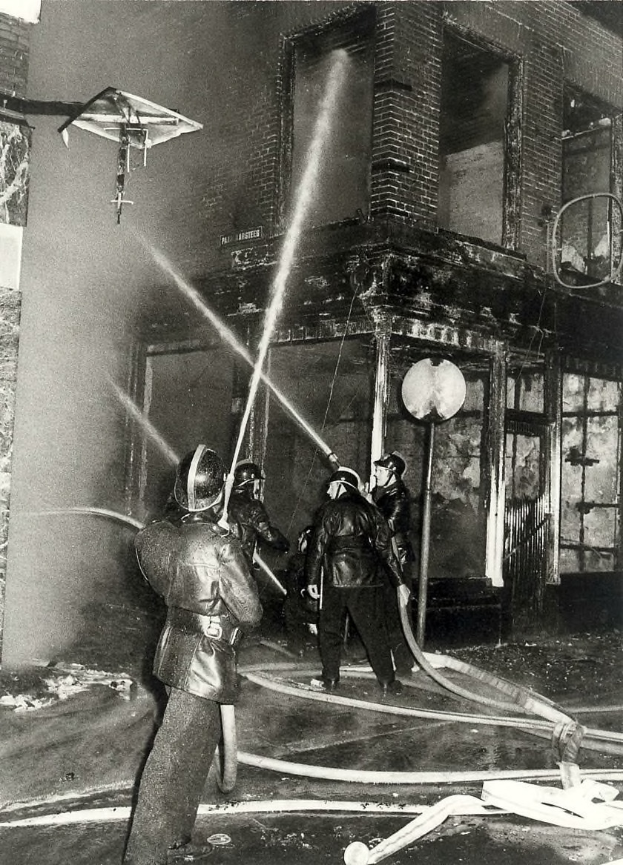 Eine Gruppe von Feuerwehrmännern in Helmen, die Rohre halten, steht vor einem feuerbeschädigten Gebäude, mit Text unten auf dem Schwarz-Weiß-Bild.