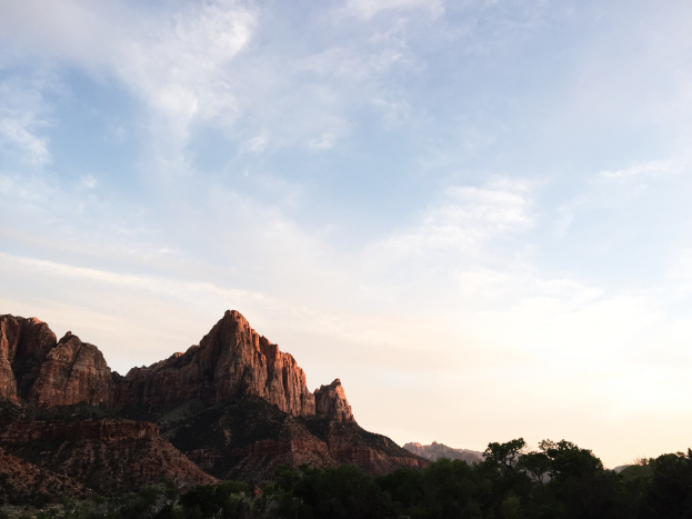 Ein malerischer Blick auf den Zion-Nationalpark in Utah mit Bäumen, Hügeln und einem Himmel mit Wolken, der von der untergehenden Sonne erhellt wird.