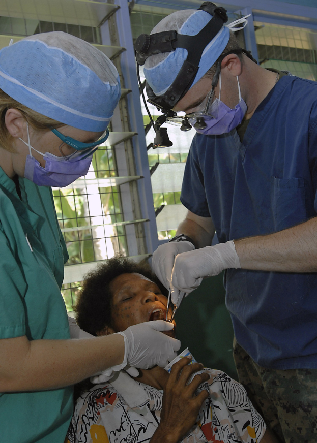 Zwei maskierte Chirurgen in Kappen und Handschuhen führen eine Gesichtsoperation an einer Frau durch, die auf einem Bett liegt, mit einem Fenster und Bäumen im Hintergrund.