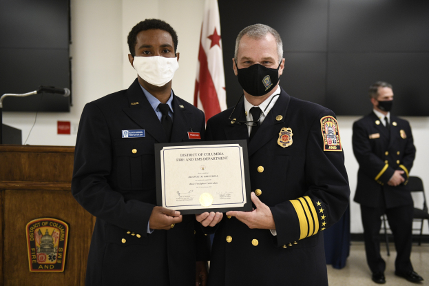 Zwei uniformierte Männer mit Masken stehen nebeneinander und halten eine Urkunde, wobei der Mann rechts die Urkunde an den Mann in der Mitte übergibt, der als District of Columbia Fire and EMS Officer of the Year identifiziert wird.