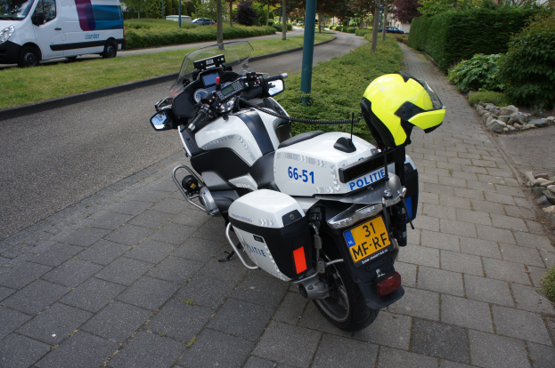 Eine Polizeimotorrad auf dem Seitenstreifen geparkt mit einem Helm darauf, umgeben von Bäumen, Pfählen, Gebäuden, Pflanzen, Gras, Steinen und anderen Fahrzeugen im Hintergrund.