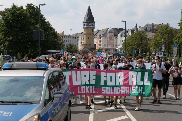 Eine große Gruppe von Menschen marschiert mit einem Banner mit der Aufschrift "Lokale Solidarität" die Straße entlang, wobei ein Polizeiwagen am Straßenrand geparkt ist und Gebäude, Bäume und Straßenelemente im Hintergrund zu sehen sind.
