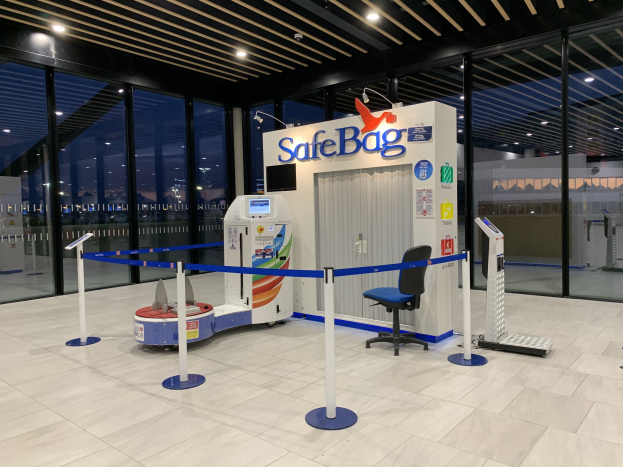 Sicherheitskontrolle am Flughafen mit einem Stuhl, Absperrgittern, Maschinen, einem Bildschirm, einer bedruckten Tafel, Glaswänden, Deckenleuchten und sichtbarem Boden.