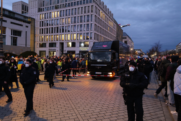 Eine Gruppe von Menschen steht vor einem Lkw auf einer Straße, umgeben von Gebäuden, Laternenmästen, Bäumen und einem bewölktem Himmel, wobei einige Mötzen und Masken tragen und ein Band mit einem Pfahl im Vordergrund ist.