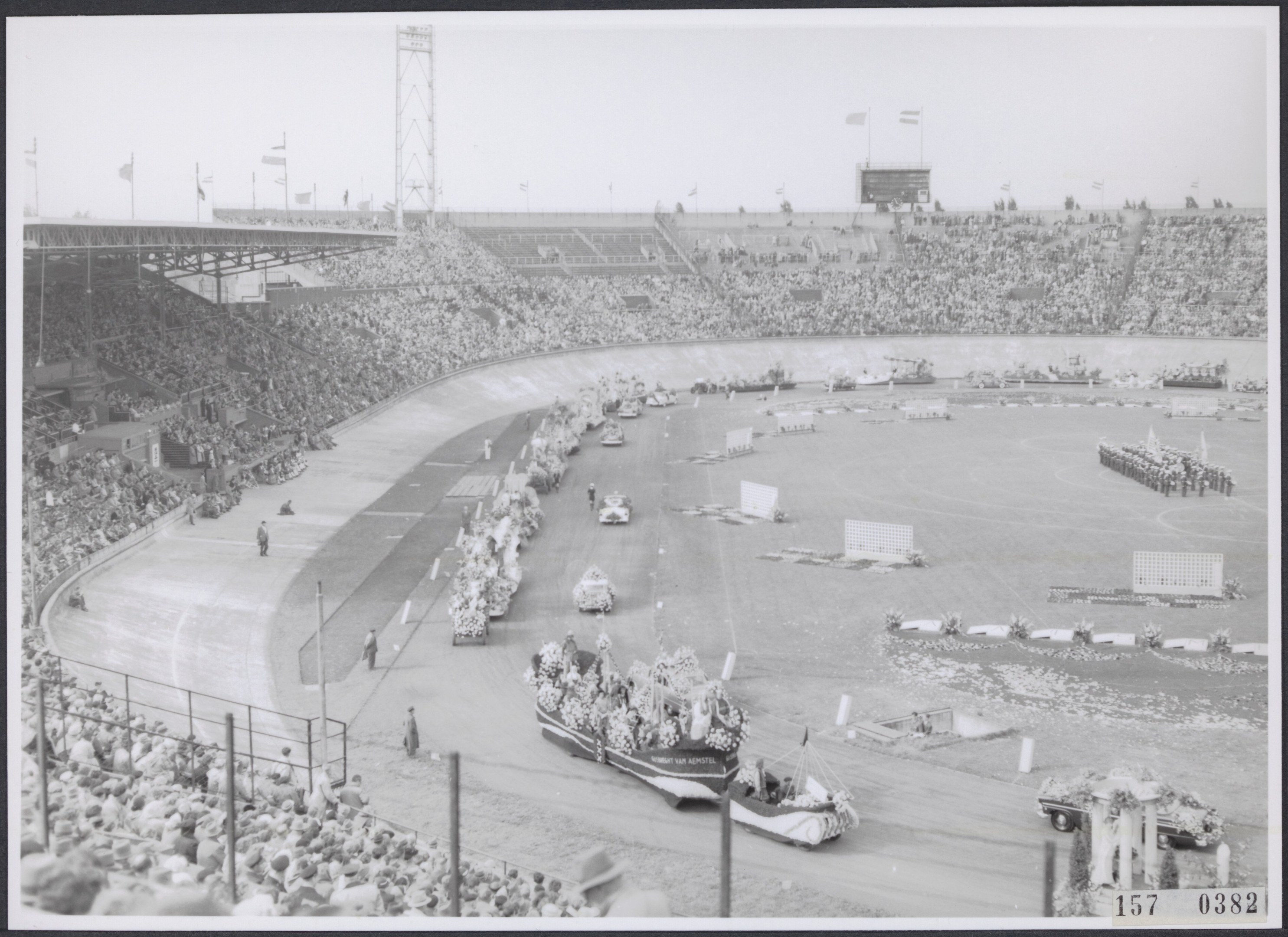 Schwarzes und weißes Foto einer Rennstrecke mit Zuschauern, die eine Gruppe von Menschen auf Fahrzeugen beobachten, einige stehen, einige sitzen, mit Pfosten, Flaggen und Himmel im Hintergrund.