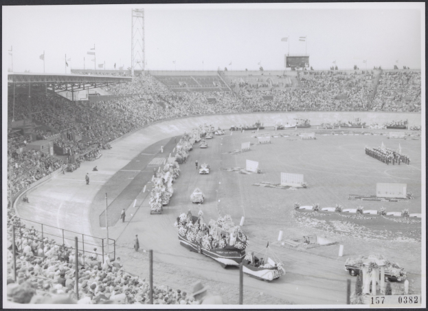 Schwarzes und weißes Foto einer Rennstrecke mit Zuschauern, die eine Gruppe von Menschen auf Fahrzeugen beobachten, einige stehen, einige sitzen, mit Pfosten, Flaggen und Himmel im Hintergrund.
