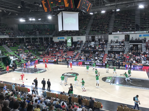 Basketballspiel in einer vollen Arena mit Zuschauern, Anzeigetafeln und Stadionbeleuchtung.