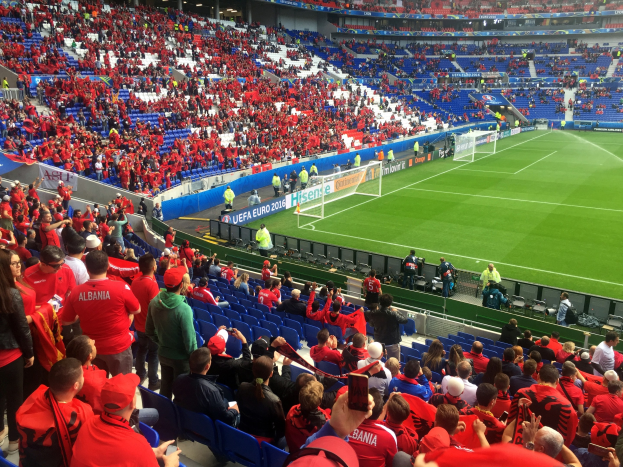 Eine große Menschenmenge in einem Stadion schaut einem Fußballspiel zu, mit Sitzplätzen und Stehplätzen, Fahnen, einem Torpfosten und Deckenbeleuchtung.