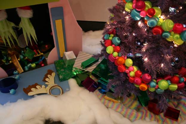 Drinnen steht ein geschmückter Weihnachtsbaum mit Lichterkette und Schmuck auf der rechten Seite, mit Geschenkboxen und Dekorationen in der Mitte, einer Hütte mit Dekorationen oben links und Baumwolle und Wolle unten.