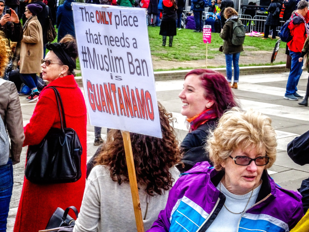 Eine Gruppe von Menschen steht auf dem Boden, einige halten Taschen, eine Frau hält ein Schild mit der Aufschrift "Der einzige Ort, an dem ein Muslimverbot gebraucht wird, ist Guantanamo" vor einem Metallzaun, Gras, einem Schild, Pfählen, einem Gebäude mit Fenstern und Bäumen.