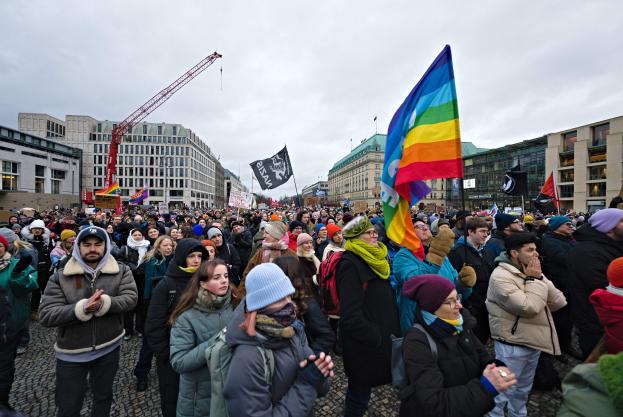 Eine große Gruppe von Menschen auf einer LGBTQ+-Rechtsmarschs in Berlin, die Fahnen und Plakate halten, mit Gebäuden, einem Kran und einem bewölkten Himmel im Hintergrund.