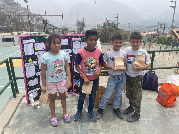 Eine Gruppe von Kindern steht auf einem Basketballfeld, jedes hält eine Papiertüte in der Hand, mit Tüten und Gegenständen rechts daneben und einer Tafel mit Text links daneben, mit Zäunen, Pfosten, Laternen, Bäumen, Gebäuden, Hügeln und Himmel im Hintergrund.