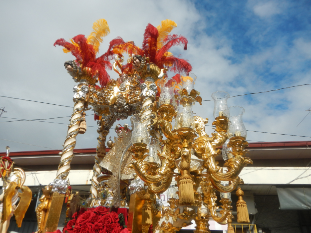 Ein prächtiger goldener und roter Wagen, geschmückt mit Blumen und anderen Ziergegenständen bei einem Karnevalsumzug, mit einem Gebäude, Strommasten mit Drähten und einem bewölkten Himmel im Hintergrund.