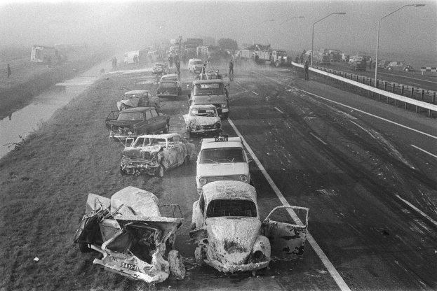 Ein Schwarz-Weiß-Foto von mehreren verunglützten Autos an der Straße bei Nacht, beleuchtet von Straßenlaternen, mit einigen Menschen in der Nähe und Bäumen und einem Gewässer im Hintergrund.