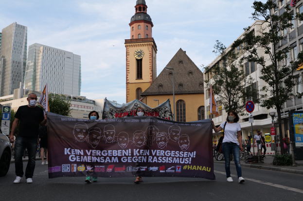 Menschen in Masken gehen eine Straße entlang und halten ein Banner hoch, mit einem geparkten Auto, Gebäuden, Bäumen, Schildern, Pfählen, einem Uhrenturm und einem klaren blauen Himmel im Hintergrund.