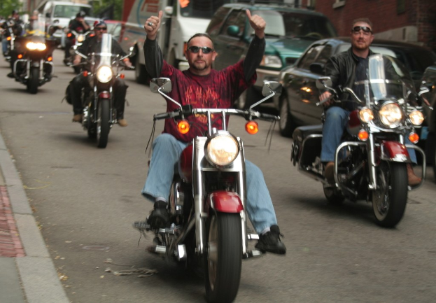 Eine Gruppe von Menschen, die auf Fahrrädern auf einer Straße fahren, wobei eine Person in einem roten Hemd ohne Hände auf dem Lenker und eine andere in einer schwarzen Jacke im Hintergrund lächelt, zusammen mit zwei Autos und einem Lieferwagen.