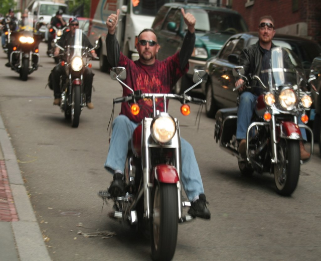 Eine Gruppe von Menschen, die auf Fahrrädern auf einer Straße fahren, wobei eine Person in einem roten Hemd ohne Hände auf dem Lenker und eine andere in einer schwarzen Jacke im Hintergrund lächelt, zusammen mit zwei Autos und einem Lieferwagen.