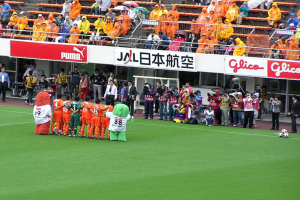 Ein Fußballspiel in einem Stadion mit sechs Spielern, drei Fußballen, vielen Zuschauern in Regenschutzjacken mit Schirmen und mehreren Kameramännern.