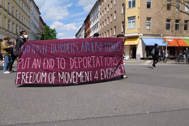 Eine Gruppe von Menschen marschiert mit einem Transparent, auf dem "Abolish Borders, Abolish Frontiers, Put an End to Deportations, Freedom of Movement 4 Everyone" steht, eine Straße entlang mit Gebäuden, Bäumen, Fahrrädern und einem bewölkten Himmel im Hintergrund.