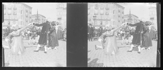 Ein Schwarz-Weiß-Foto einer Gruppe von Menschen, die auf der Straße tanzen, während eines Faschingsumzugs, gekleidet in Kostümen, mit Gebäuden, Straßenlaternen und Straßenlaternen im Hintergrund unter einem klaren Himmel.