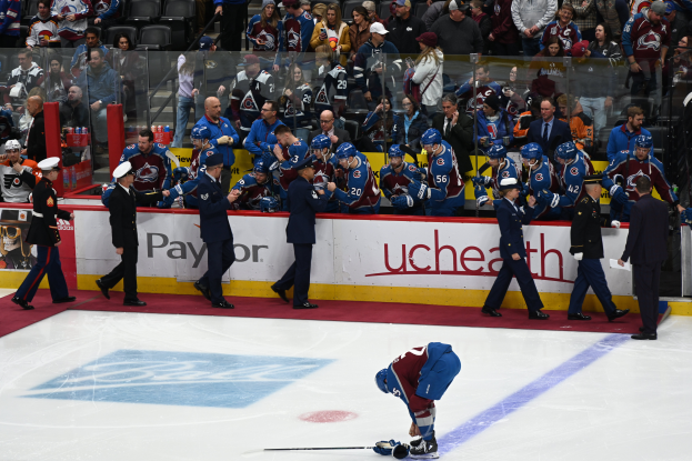 Ein Hockey-Spieler auf dem Eis mit einem Stock in der Hand, umgeben von anderen Spielern mit Stöcken und Mützen, mit Zuschauern und Brettern mit der Aufschrift "Colorado Avalanche vs San Jose Sharks - 3/24/15 NHL Pick, Odds, and Prediction" im Hintergrund.