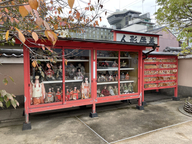 Ein kleiner Straßenaltar mit Puppen in Glaskästen, umgeben von Gebäuden, Pfählen und Drähten, mit einem roten Gebäude und einem Baum im Hintergrund.