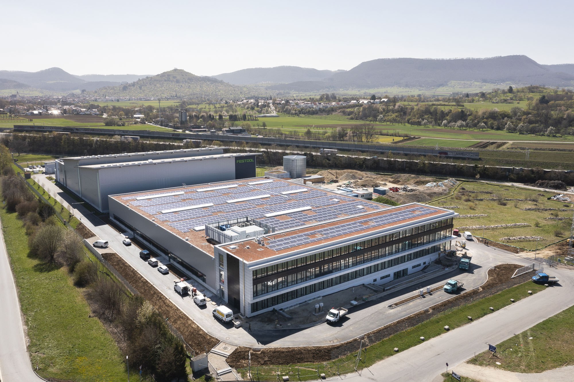 Luftaufnahme eines große Gebäudes mit Solarmodulen auf dem Dach, umgeben von Fahrzeugen, Bäumen, Gras und Strommasten, vor Hühgeland und einem klaren blauen Himmel; das Gebäude ist die erste solarbetriebene Fabrik Deutschlands.
