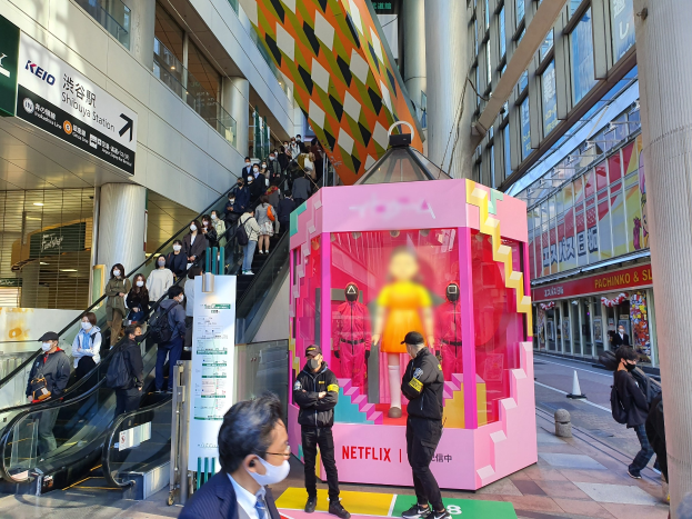 Eine Gruppe von Menschen steht vor einem Gebäude mit Glaswänden, Säulen und Texttafeln, einige tragen Masken, während eine Rolltreppe mit Menschen zu sehen ist. In der Mitte befindet sich eine pinkfarbene Box mit der Aufschrift "Netflix's new Netflix booth in Tokyo", die Puppen enthält.