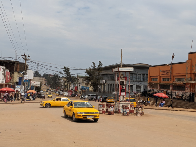 Ein gelbes Taxi fährt eine belebte Stadtstraße entlang, gesäumt von Gebäuden, während Fußgänger mit Regenschirmen auf dem Gehweg darunter gehen, mit Strommasten und Drähten im Hintergrund.