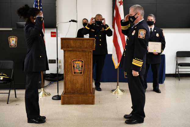 Gruppe uniformierter Personen in Masken vor einem Podium mit Mikrofon, umgeben von Flaggen, Stühlen und Tischen, bei einer Vereidigung eines neuen Feuerwehrchefs.