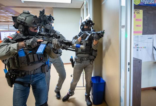 Eine Gruppe von Polizisten in Uniform, die Helme tragen und Waffen halten, steht in einem Raum mit einem Müllcontainer, einer Tafel mit Papieren, einer Tür, einem Monitor und Deckenleuchten.