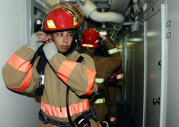 Feuerwehrleute in Uniform, die während einer Übung in einem Raum stehen, mit Rohren und Equipment im Hintergrund.