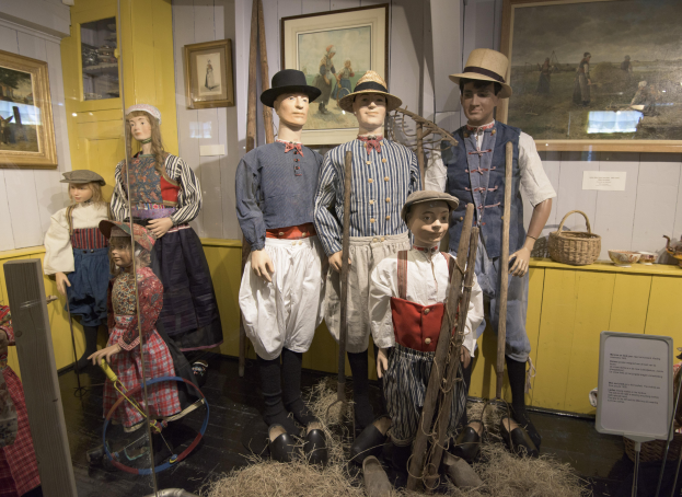 Eine Gruppe von Schaufensterpuppen in historischer Kleidung steht zusammen in einem Raum mit trockenem Gras auf dem Boden, einem Textbrett, gerahmten Fotos an den W├Ąnden, einem Schrank mit Gegenst├Ąnden und einem Korb und anderen Gegenst├Ąnden auf einer Plattform, wahrscheinlich im Nationalen Historischen Museum der Vereinigten Staaten.