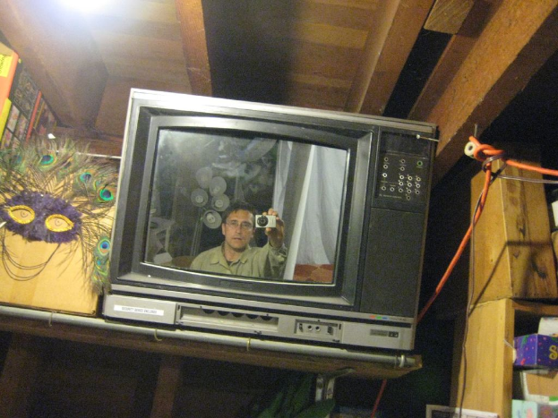 Ein Fernsehbildschirm mit einer Person umgeben von Pfauenfedern, BÃ¼chern und Schachteln.