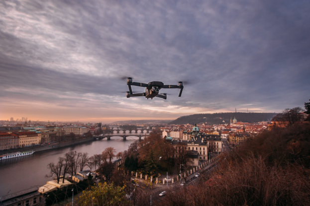 Eine Drohne fliegt über einer Stadt mit Gebäuden, Bäumen und Fahrzeugen, mit einem Fluss und einer Brücke im Hintergrund.