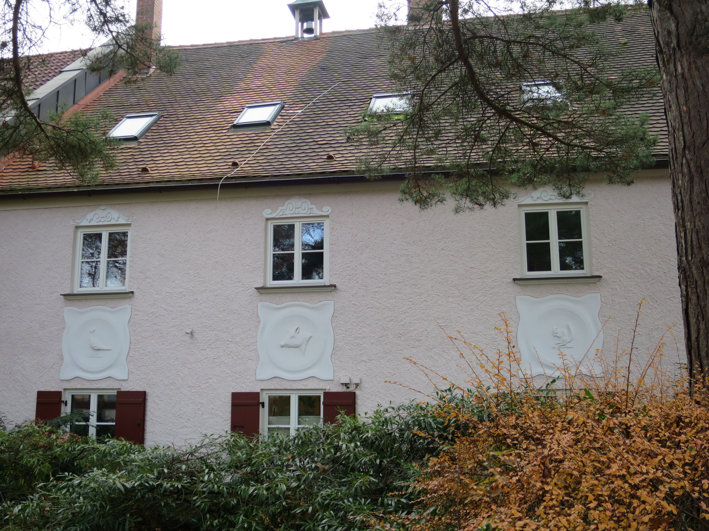 Ein weißes Haus mit roten Fensterläden, mehreren Fenstern und einem Baum davor, umgeben von grünen Pflanzen unter einem hellblauen Himmel.