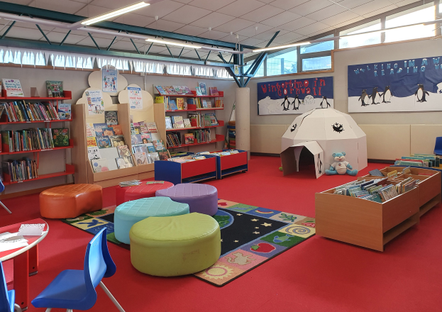 Eine hell erleuchtete Kinderbibliothek mit Bücherregalen, Tischen, Stühlen, Hockern, Teppichen, Wanddisplays, Deckenlampen und großen Fenstern.