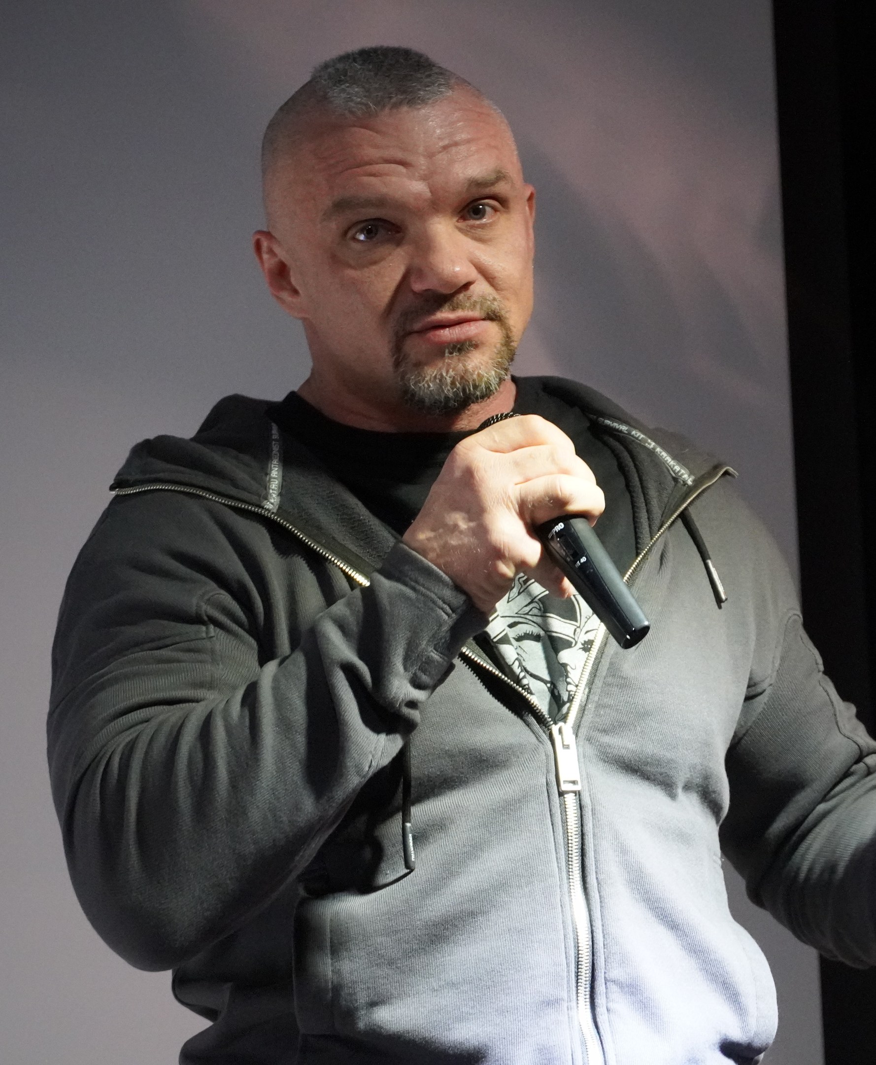 Ein Mann in einer schwarzen Kapuzenjacke und einem schwarzen Anzug mit einem weißen Hemd hält ein Mikrofon und steht selbstbewusst vor einer Wand.