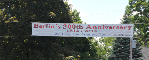 Ein Banner mit der Aufschrift "Berliner Jubiläum 200" hängt an einem Seil im Vordergrund, mit Bäumen im Hintergrund und einem Haus auf der rechten Seite, unter einem sichtbaren Himmel.