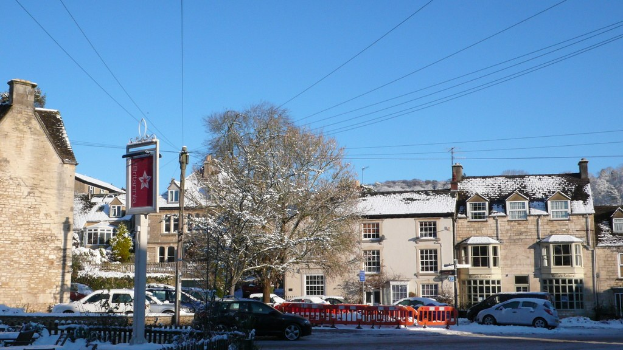 Fahrzeuge auf einer schneebedeckten Straße mit Häusern, Bäumen, Strommasten und schneebedeckten Drähten im Hintergrund.