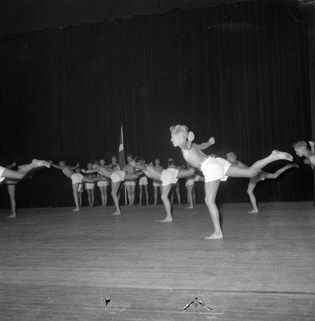 Schwarzes Gruppenfoto von Turnern auf einer Bühne mit Vorhang und Flaggenhintergrund.