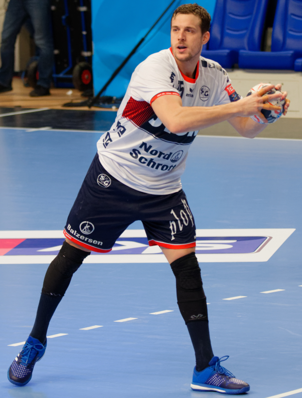 Ein Mann in einem weißen Hemd und blauen Shorts spielt Handball, hält einen Ball und hat im Hintergrund Stühle, eine stehende Person, eine Tribüne und andere Gegenstände.