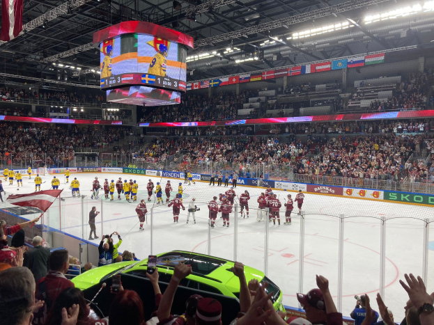 Ein Eishockeyspiel in einer großen Arena mit Zuschauern, einige halten Fahnen, ein Auto davor, Bande, Banner, Deckenbeleuchtung, ein Bildschirm und hängende Fahnen.